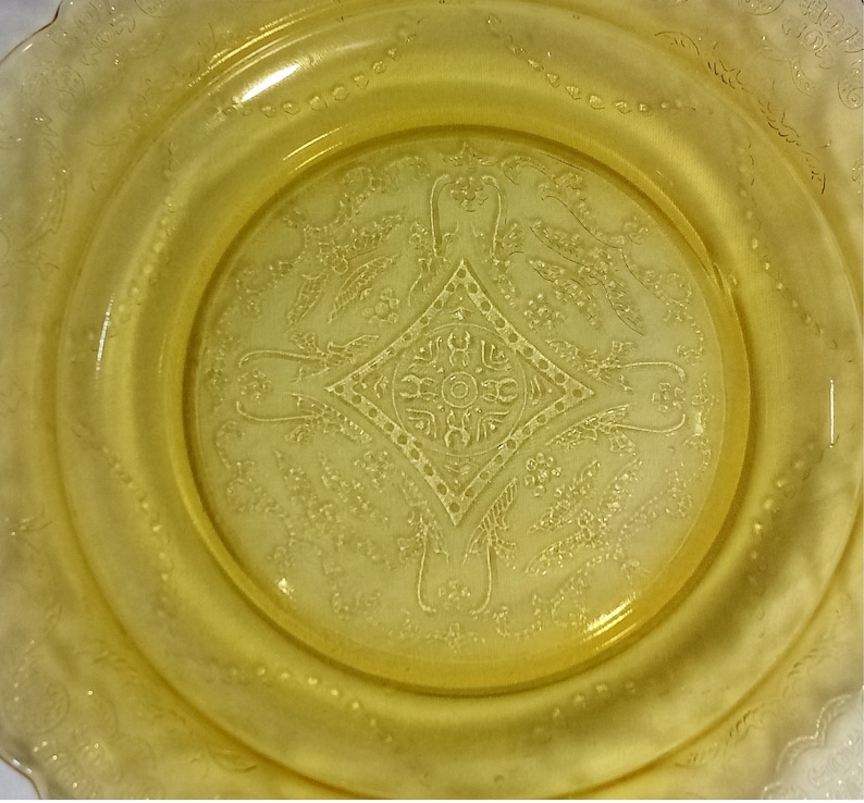 Pu&ograve; includere: Un piatto di vetro giallo con un motivo decorativo di fiori e foglie. Il piatto ha un bordo ondulato e un centro rialzato.