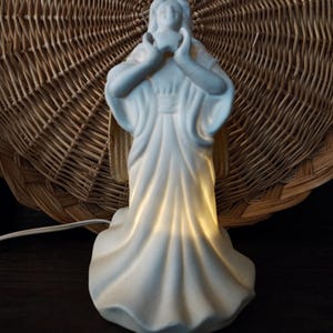 Puede incluir: Una figura de cerámica blanca de una mujer con un vestido fluido, con las manos juntas cerca de su rostro. La figura está iluminada desde dentro, creando un brillo suave. El fondo presenta una cesta tejida.