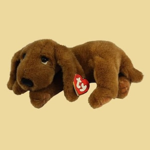 Puede incluir: Un perro de peluche marrón en posición tumbada con orejas largas y caídas, nariz negra y ojos oscuros. Una etiqueta roja en forma de corazón con las letras blancas ty está sujeta al costado del peluche cerca de la pata delantera.