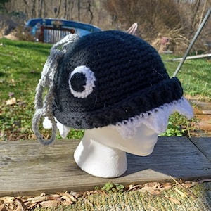 May include: A handmade crochet hat in the shape of a cartoon character. The hat is primarily black with white and gray accents, including a large eye and dangling gray yarn. The hat is displayed on a white mannequin head.