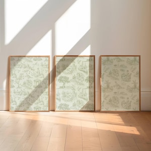 May include: Three framed prints with a light green and white toile pattern, leaning against a white wall. The frames are a light brown color. The room has a wooden floor and natural light from a window.