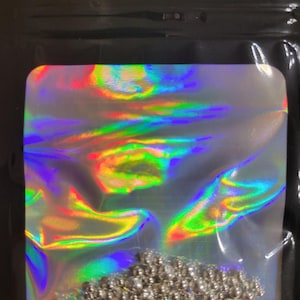 May include: A clear, holographic bag containing a collection of small, silver-colored beads. The bag reflects a spectrum of colors, including red, yellow, and blue. The beads are clustered at the bottom of the bag.