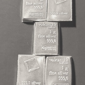 May include: A collection of six silver bars, each stamped with "valcambi suisse", "1 g fine silver 999.0", and a logo. The bars are rectangular with a slightly textured surface, arranged on a dark background.