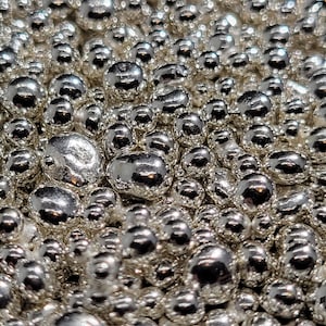 May include: A close-up shot of numerous shiny, silver-coloured metal beads. The beads are of varying sizes and reflect light, creating a textured, reflective surface. The overall effect is a dense, metallic display.