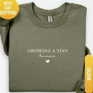 May include: Olive green sweatshirt with the text "GROWING A TINY human" in white, along with a small white heart. The sweatshirt is a Gildan brand and is labeled "MILITARY GREEN".