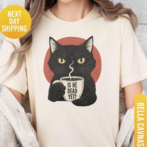 Może przedstawiać: Kremowa koszulka z czarnym kotem trzymającym kubek z kawą. Na kubku napis "IS HE DEAD YET?". Kot jest umieszczony na czerwonym okręgu. Koszulka jest oznaczona jako "SOFTCREAM" i "BELLA+CANVAS".