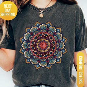May include: Dark grey t-shirt featuring a colorful mandala design with the letters "FDT" and "EBI". The mandala is in shades of blue, yellow, and red. The shirt has short sleeves and a crew neck. The text "Comfort Colors" and "Pepper" are also visible.