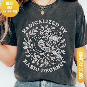May include: Dark grey t-shirt with a white graphic of a bird surrounded by flowers and leaves, with the text "RADICALIZED BY BASIC DECENCY". The shirt has a round neck and short sleeves. The shirt is labeled "Pepper" and "Comfort Colors".