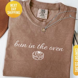 May include: Espresso-colored t-shirt with the phrase "bun in the oven" and a graphic of a cinnamon roll. A silver necklace with a pendant rests on the shirt. The shirt is folded and displayed with a hat and dried plant.