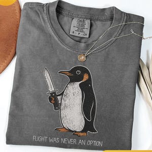May include: A charcoal grey t-shirt featuring a graphic of a penguin holding a serrated knife. Below the penguin, the text reads, "FLIGHT WAS NEVER AN OPTION." The shirt has a crew neckline and a Comfort Colors brand label printed on the inside neck.