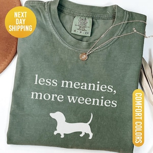May include: Moss green Comfort Colors t-shirt with the text "less meanies, more weenies" and a white dachshund graphic. A gold necklace with a pendant is on the shirt. The shirt has a crew neck and short sleeves.