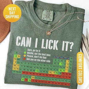 May include: Moss green Comfort Colors t-shirt with the text "CAN I LICK IT?" and a periodic table design. The shirt has a crew neck and short sleeves. A gold necklace with a pendant is on the shirt.
