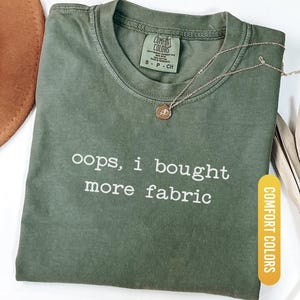 Könnte beinhalten: Ein moosgrünes T-Shirt mit dem weißen Text "oops, i bought more fabric". Das Shirt hat einen Rundhalsausschnitt und kurze Ärmel. Eine goldene Halskette mit einem kleinen Anhänger liegt auf dem Shirt. Das Shirt ist mit "Comfort Colors" gekennzeichnet.