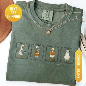 May include: Moss green Comfort Colors t-shirt featuring a graphic of four ducks reading books, each in a framed illustration. A gold necklace with a pendant rests on the shirt. The t-shirt has a crew neck and short sleeves.