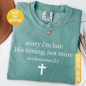 May include: Light green t-shirt with the text "sorry i'm late His timing, not mine ecclesiastes 3:1" and a white cross. The shirt has a crew neck and short sleeves. A gold necklace with a pendant is on the shirt.