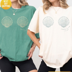 May include: Two short-sleeve crewneck t-shirts, one in light green and one in ivory, each featuring a graphic of two seashells on the chest. Below the right shell, the text reads Cabo San Lucas June 2025. Both shirts are paired with black shorts.