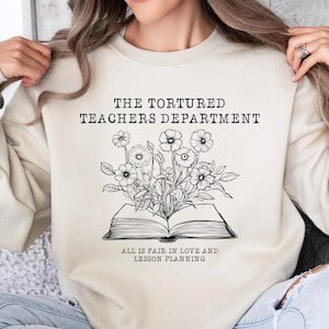 May include: A cream-coloured sweatshirt with the text "The Tortured Teachers Department" and a black and white illustration of a book with flowers growing out of it. The text below the book reads "All is fair in love and lesson planning."