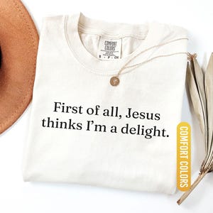 May include: Ivory-colored t-shirt with the text "First of all, Jesus thinks I'm a delight." A gold-colored necklace with a pendant is on the shirt. The shirt has a Comfort Colors tag.