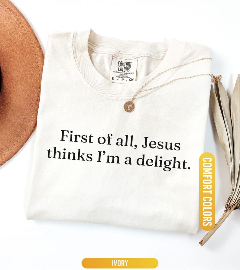 May include: Ivory-colored t-shirt with the text "First of all, Jesus thinks I'm a delight." A gold-colored necklace with a pendant is on the shirt. The shirt has a Comfort Colors tag.