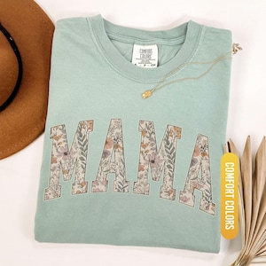 Op de afbeelding: Een lichtgroen t-shirt met het woord "MAMA" in een bloemenpatroon. Het shirt heeft een ronde hals en korte mouwen. Een gouden ketting en een bruine hoed zijn ook zichtbaar.