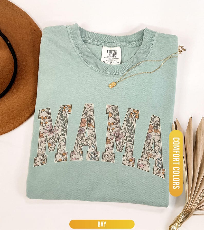 Op de afbeelding: Een lichtgroen t-shirt met het woord "MAMA" in een bloemenpatroon. Het shirt heeft een ronde hals en korte mouwen. Een gouden ketting en een bruine hoed zijn ook zichtbaar.