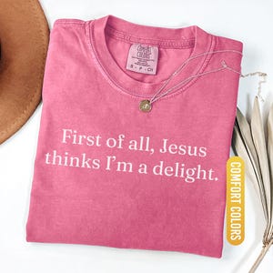 May include: A pink Comfort Colors t-shirt with the text "First of all, Jesus thinks I'm a delight." A silver necklace with a pendant is on the shirt. The shirt is labeled "Crunchberry."