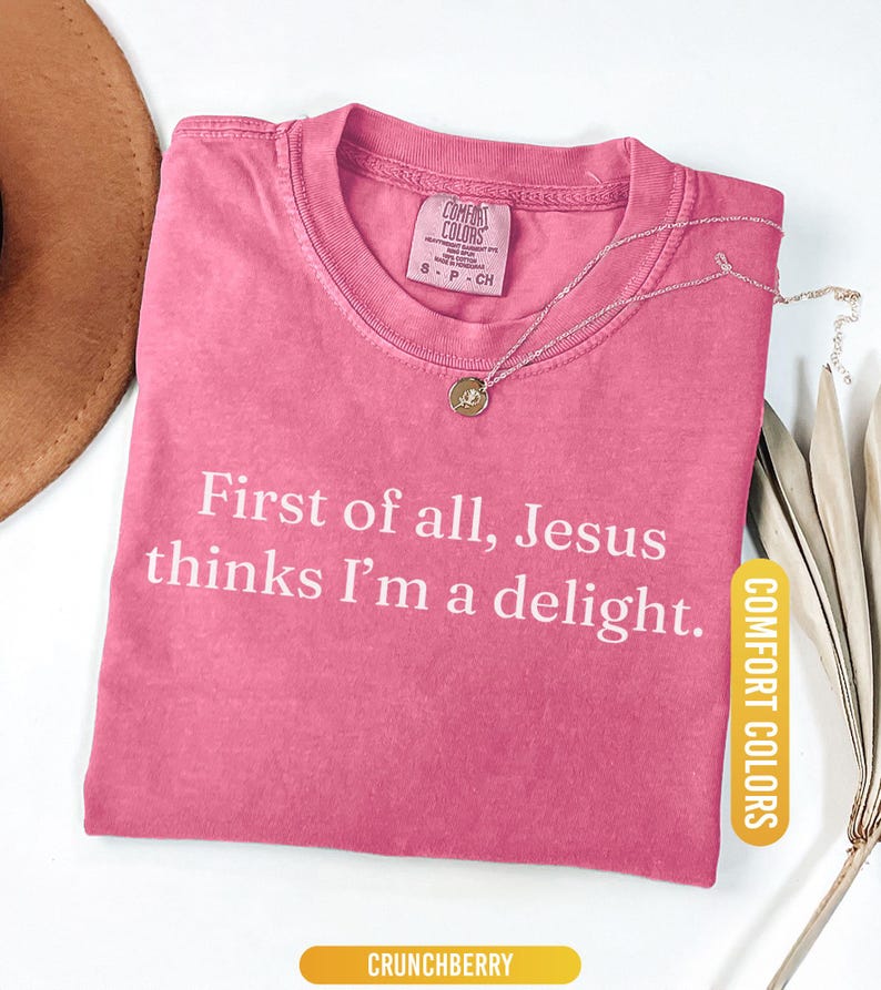 May include: A pink Comfort Colors t-shirt with the text "First of all, Jesus thinks I'm a delight." A silver necklace with a pendant is on the shirt. The shirt is labeled "Crunchberry."