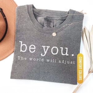 May include: A gray Comfort Colors t-shirt with the words "be you. The world will adjust" printed in white. A gold necklace with a circular pendant rests on the shirt. The shirt is labeled "Pepper".
