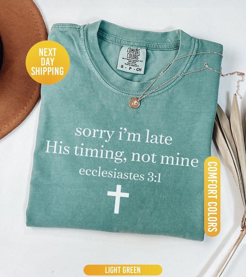 May include: Light green t-shirt with the text "sorry i'm late His timing, not mine ecclesiastes 3:1" and a white cross. The shirt has a crew neck and short sleeves. A gold necklace with a pendant is on the shirt.