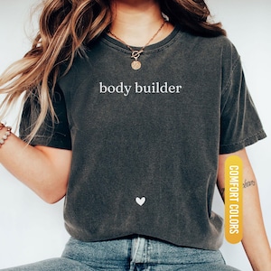 Pu&ograve; includere: T-shirt grigio scuro con la scritta bianca "body builder". Un piccolo cuore bianco &egrave; sotto il testo. La maglietta &egrave; etichettata "Comfort Colors" e "Pepper".