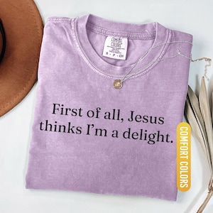 May include: Orchid-colored t-shirt with the text "First of all, Jesus thinks I'm a delight." A silver necklace with a pendant rests on the shirt. The shirt has a Comfort Colors tag. The word "ORCHID" is at the bottom.
