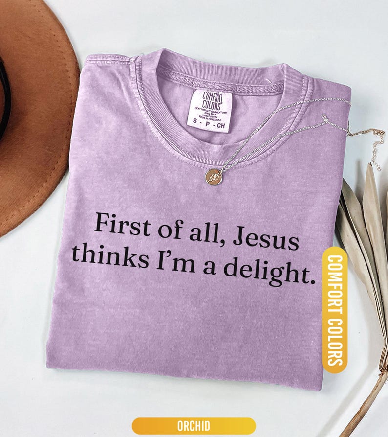 May include: Orchid-colored t-shirt with the text "First of all, Jesus thinks I'm a delight." A silver necklace with a pendant rests on the shirt. The shirt has a Comfort Colors tag. The word "ORCHID" is at the bottom.