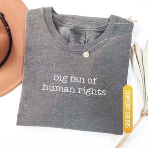 May include: A gray Comfort Colors t-shirt with the embroidered text "big fan of human rights." A gold necklace with a circular pendant rests on the shirt. A brown hat with a black strap is visible in the background.