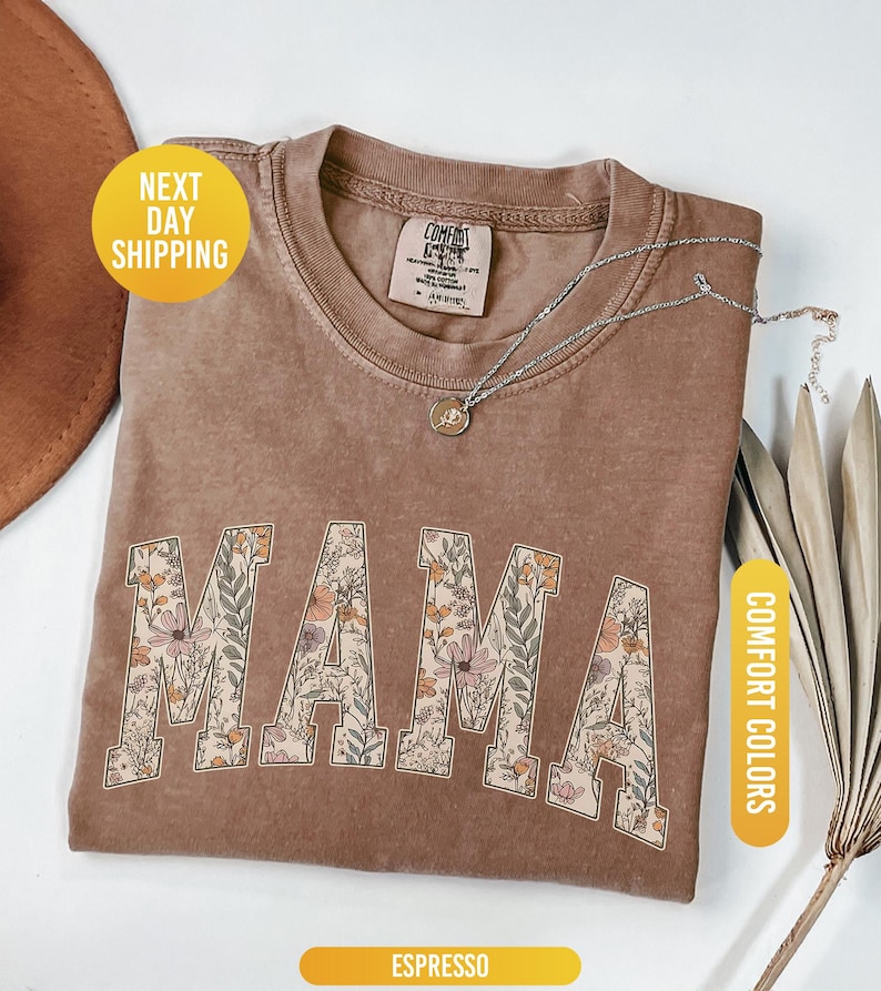 Op de afbeelding: Espresso-kleurig t-shirt met het woord "MAMA" in een bloemenontwerp. Het shirt heeft een ronde hals en korte mouwen. Een zilveren ketting met een hanger is over het shirt gedrapeerd. Het shirt is gelabeld "Comfort Colors".