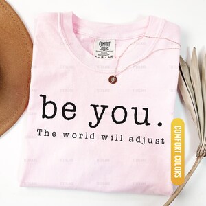 May include: A light pink t-shirt with the words "be you. The world will adjust" in black. A rose gold necklace and a brown hat are also visible. The shirt is labeled "Blossom" and "Comfort Colors."