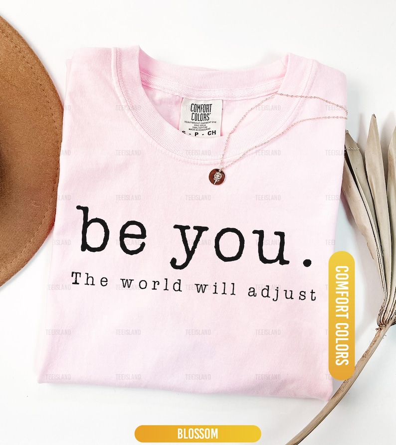 May include: A light pink t-shirt with the words "be you. The world will adjust" in black. A rose gold necklace and a brown hat are also visible. The shirt is labeled "Blossom" and "Comfort Colors."