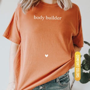 Pu&ograve; includere: T-shirt arancione bruciato con la scritta bianca "body builder", un piccolo cuore bianco e le parole "Comfort Colors" su un'etichetta gialla. La maglietta &egrave; abbinata a jeans blu.