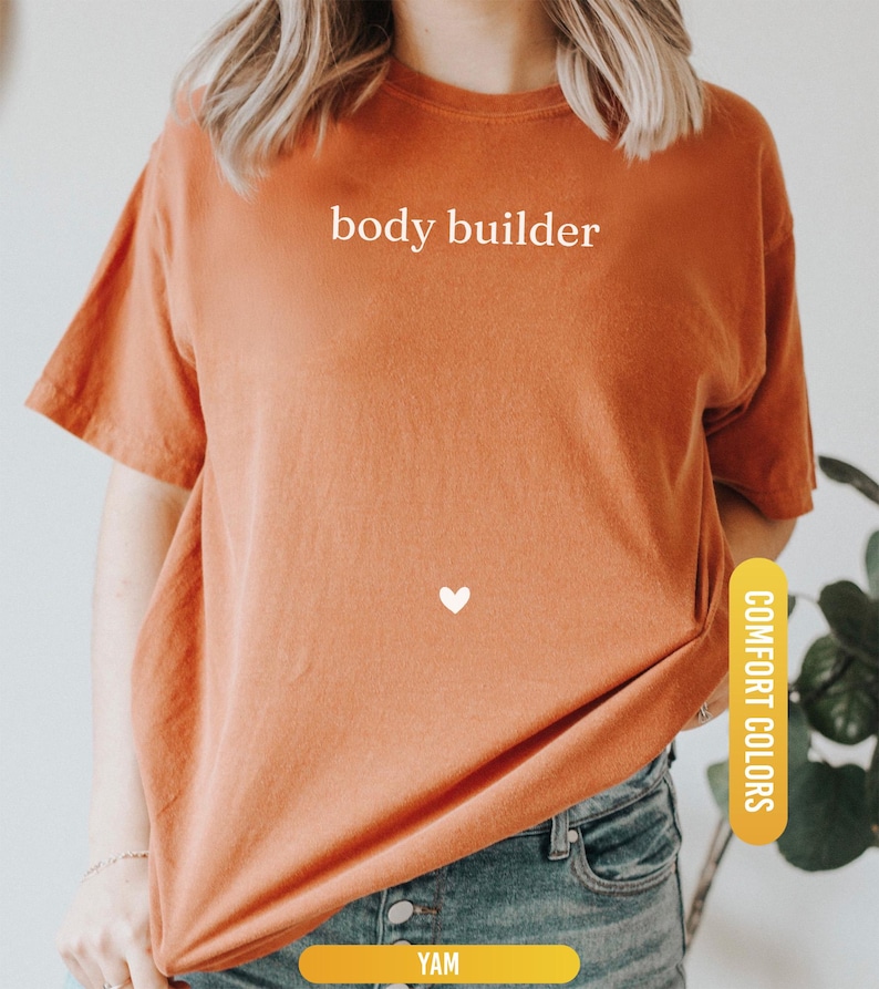 Pu&ograve; includere: T-shirt arancione bruciato con la scritta bianca "body builder", un piccolo cuore bianco e le parole "Comfort Colors" su un'etichetta gialla. La maglietta &egrave; abbinata a jeans blu.