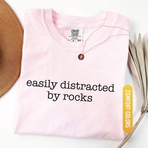 May include: A light pink t-shirt with the text "easily distracted by rocks" in black. The shirt has a crew neck and a Comfort Colors tag. A gold necklace with a pendant is on the shirt. The word "Blossom" is at the bottom.