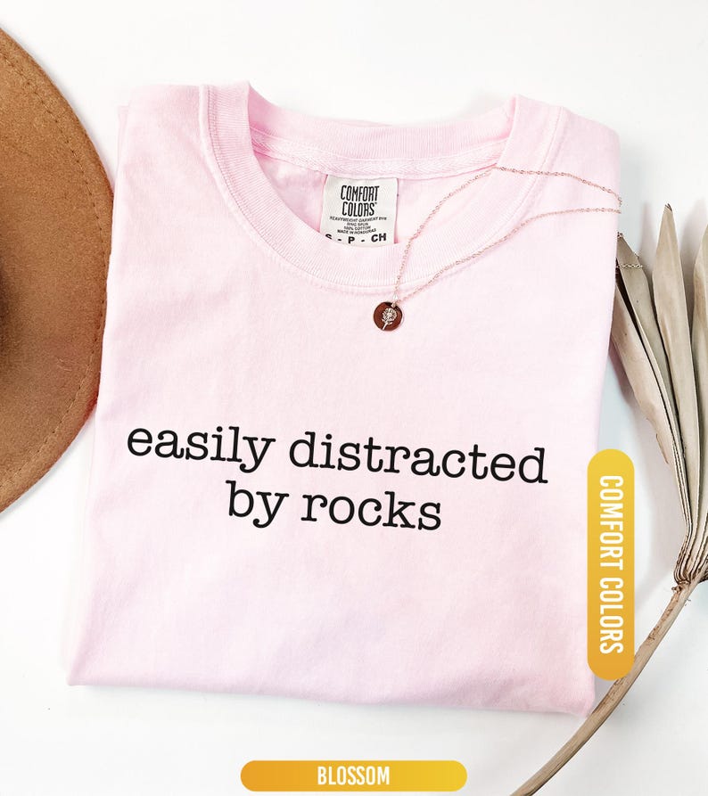 May include: A light pink t-shirt with the text "easily distracted by rocks" in black. The shirt has a crew neck and a Comfort Colors tag. A gold necklace with a pendant is on the shirt. The word "Blossom" is at the bottom.