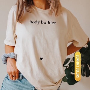 Pu&ograve; includere: T-shirt color avorio con la scritta "body builder" in nero e un piccolo cuore nero. La maglietta fa parte della collezione "Comfort Colors". La parte inferiore della maglietta ha un'etichetta gialla con la scritta "IVORY".