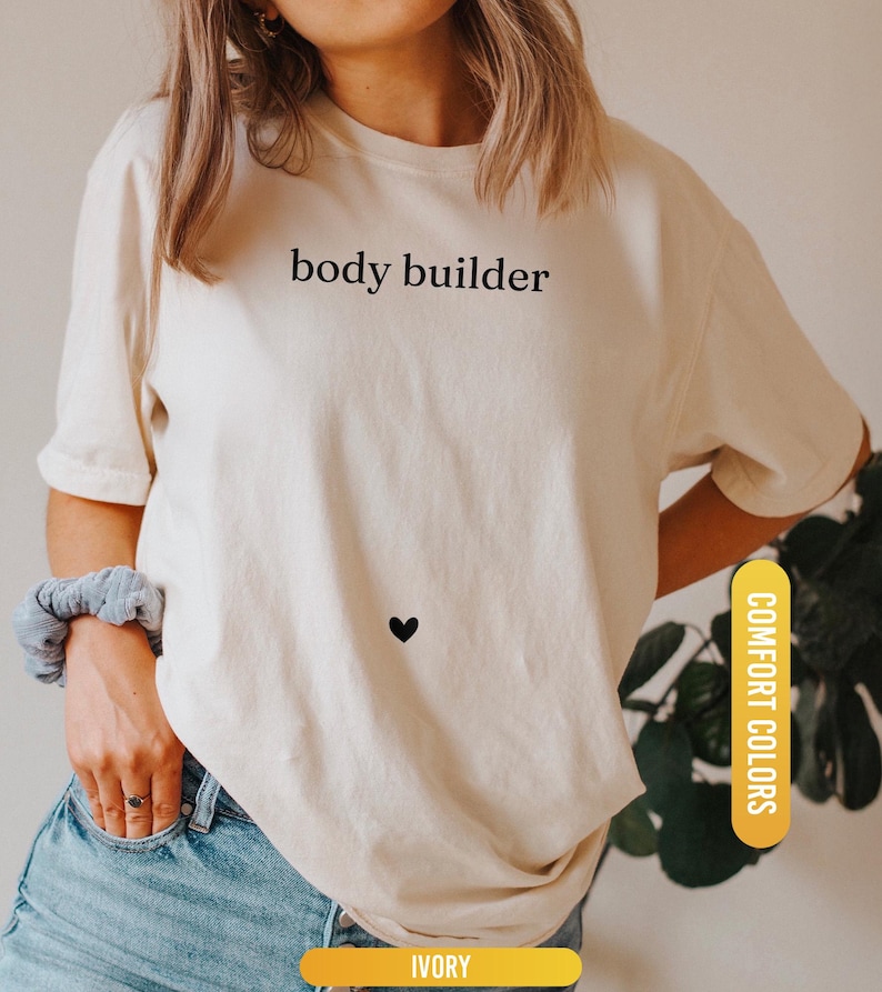 Pu&ograve; includere: T-shirt color avorio con la scritta "body builder" in nero e un piccolo cuore nero. La maglietta fa parte della collezione "Comfort Colors". La parte inferiore della maglietta ha un'etichetta gialla con la scritta "IVORY".