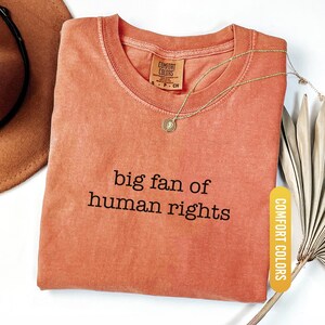 May include: A coral-colored t-shirt with the phrase "big fan of human rights" printed in black. A gold necklace with a pendant rests on the shirt. The shirt has a Comfort Colors tag.