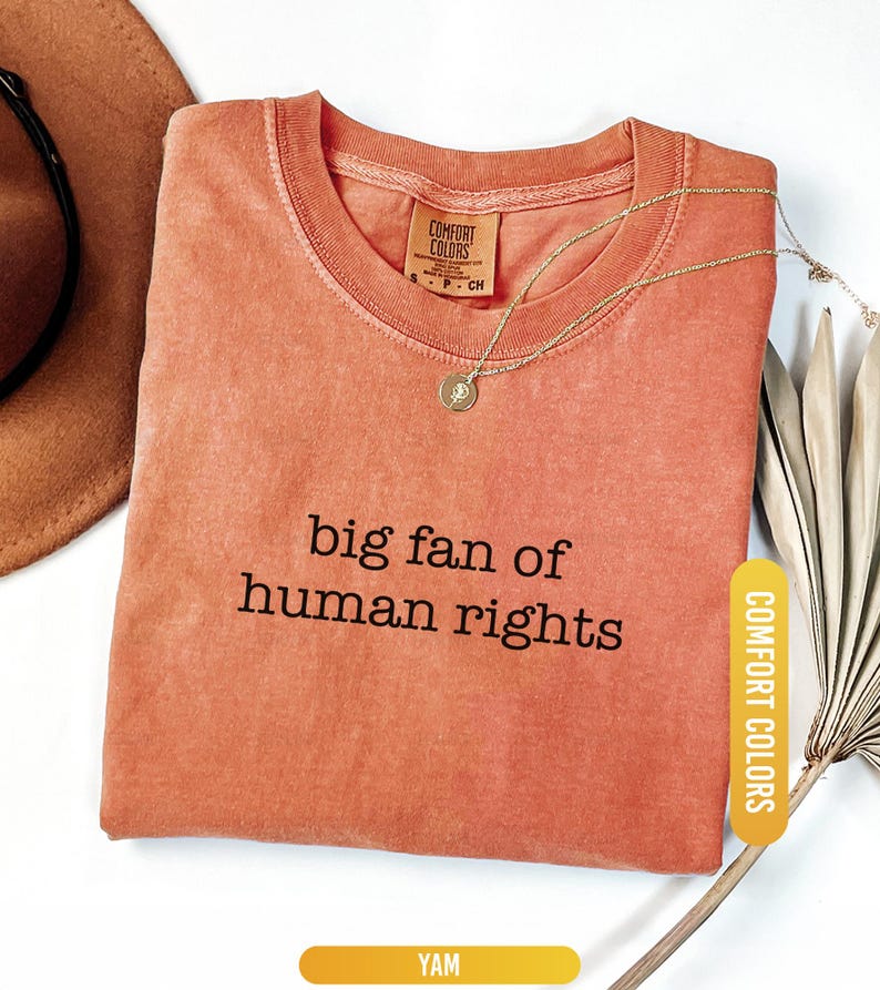 May include: A coral-colored t-shirt with the phrase "big fan of human rights" printed in black. A gold necklace with a pendant rests on the shirt. The shirt has a Comfort Colors tag.