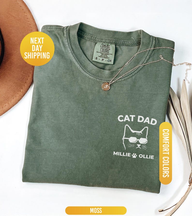 May include: A moss green t-shirt with the words "CAT DAD" and a cat graphic wearing sunglasses. The names "MILLIE & OLLIE" are printed below the cat. The shirt has a crew neck and short sleeves. The words "COMFORT COLORS" are printed on the shirt.