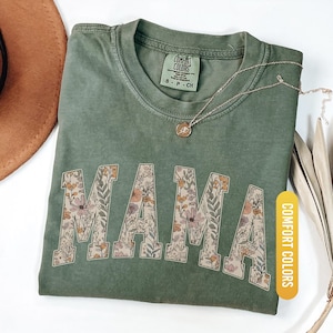 Op de afbeelding: Een mosgroen t-shirt met het woord "MAMA" in een bloemenontwerp. Het shirt heeft een ronde hals en korte mouwen. Een gouden ketting met een hanger rust op het shirt. Het shirt is gelabeld "Comfort Colors" en de kleur is "Moss".