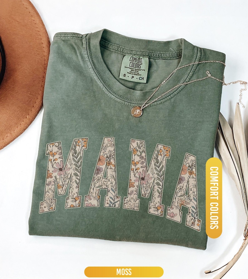 Op de afbeelding: Een mosgroen t-shirt met het woord "MAMA" in een bloemenontwerp. Het shirt heeft een ronde hals en korte mouwen. Een gouden ketting met een hanger rust op het shirt. Het shirt is gelabeld "Comfort Colors" en de kleur is "Moss".