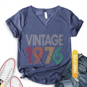May include: Heather navy V-neck t-shirt with the text "VINTAGE 1976" in a retro design. The numbers are in red, orange, yellow, and green. The shirt is paired with white sneakers and blue jeans.