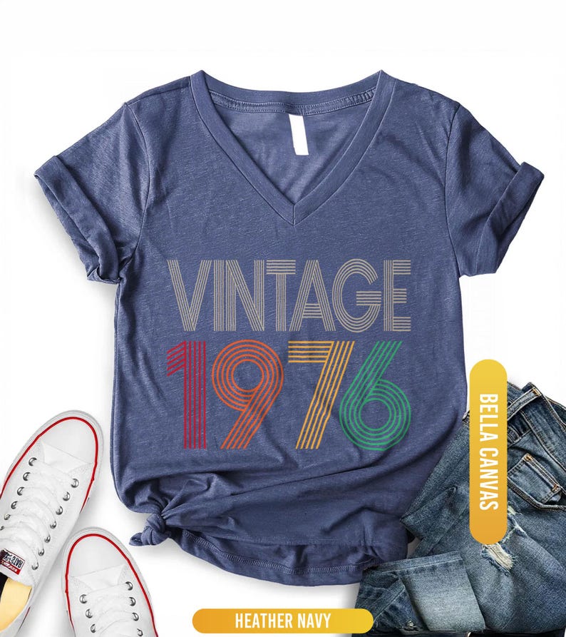 May include: Heather navy V-neck t-shirt with the text "VINTAGE 1976" in a retro design. The numbers are in red, orange, yellow, and green. The shirt is paired with white sneakers and blue jeans.