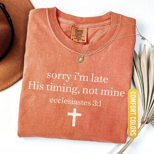 May include: A yam-colored t-shirt with the text "sorry i'm late His timing, not mine ecclesiastes 3:1" and a white cross. A gold necklace and a brown hat are also visible.